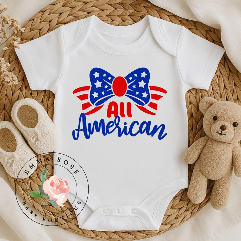 May include: White baby onesie with a patriotic design. The design features a blue bow with white stars and red stripes, with the words "All American" in blue script. A teddy bear and baby shoes are next to the onesie.