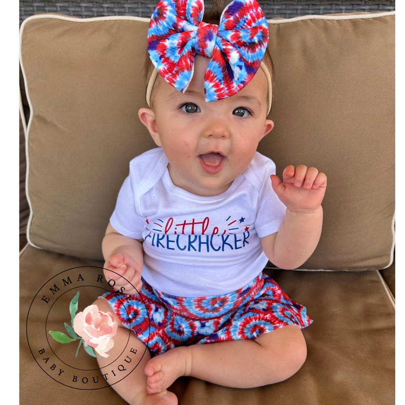 May include: A white short-sleeved bodysuit with the text little firecracker in red and blue, paired with red, white, and blue tie-dye shorts and a matching oversized tie-dye hair bow. The outfit features a patriotic color scheme throughout.