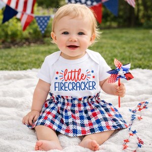 May include: A white short-sleeved bodysuit with the red and blue text "little FIRECRACKER" and a matching skirt featuring a red, white, and blue plaid pattern. The outfit is paired with a small red, white, and blue pinwheel toy.