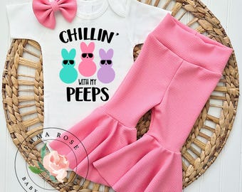 May include: A baby outfit set featuring a white onesie with the text "Chillin' with my Peeps" and three cartoon bunny figures wearing sunglasses. The set includes pink bell-bottom pants and a matching pink bow headband. The outfit is displayed on a woven basket.