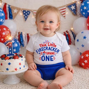 May include: A baby wearing a white onesie with the text "This Little Firecracker is Turning ONE" in red and blue. The baby is sitting in front of a cake with a number one candle and balloons.