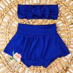 May include: A royal blue baby outfit set, including a headband with a bow and a ruffled skirted bottom. The set is made of ribbed fabric. The logo "EMMA ROSE BABY BOUTIQUE" is visible on the bottom left.