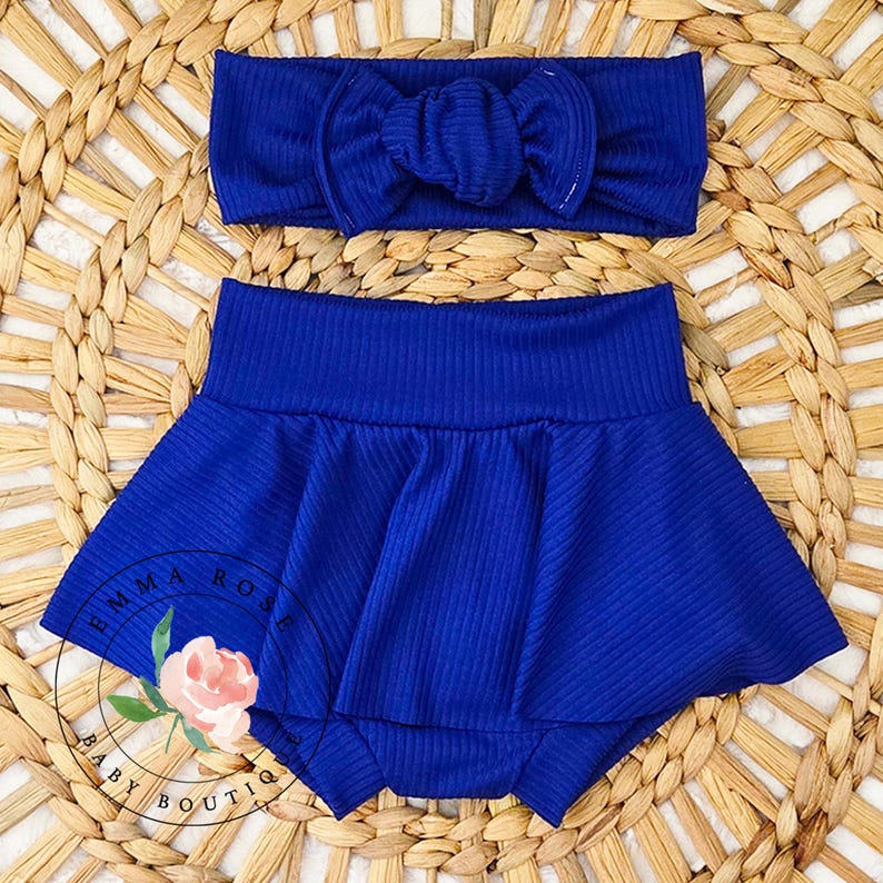 May include: A royal blue baby outfit set, including a headband with a bow and a ruffled skirted bottom. The set is made of ribbed fabric. The logo "EMMA ROSE BABY BOUTIQUE" is visible on the bottom left.