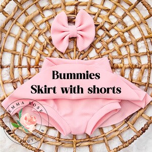 May include: A light pink textured fabric set consisting of a skirt with integrated shorts and a matching hair bow. The text Bummies Skirt with shorts is printed in black across the center. A circular logo with a floral graphic and the text Emma Rose Boutique is visible in the lower left corner.