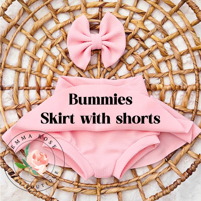 May include: A light pink textured fabric set consisting of a skirt with integrated shorts and a matching hair bow. The text Bummies Skirt with shorts is printed in black across the center. A circular logo with a floral graphic and the text Emma Rose Boutique is visible in the lower left corner.