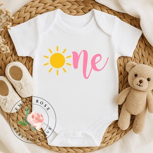 May include: A white short-sleeved baby bodysuit featuring a yellow sun graphic next to the word one written in pink cursive script. The garment has a round neckline and three snap closures at the bottom for easy access during diaper changes.