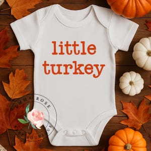 Pu&ograve; includere: Un body bianco a maniche corte per neonati con la scritta little turkey stampata in arancione sul petto. Il capo presenta uno scollo rotondo e tre bottoni a pressione sul fondo per la chiusura, con un logo circolare visibile.