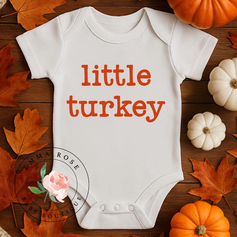 Pu&ograve; includere: Un body bianco a maniche corte per neonati con la scritta little turkey stampata in arancione sul petto. Il capo presenta uno scollo rotondo e tre bottoni a pressione sul fondo per la chiusura, con un logo circolare visibile.