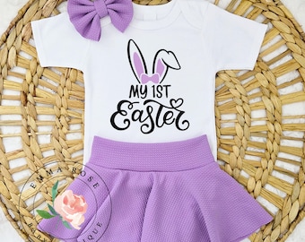 May include: A white baby bodysuit with a bunny ear graphic and the text "MY 1ST EASTER". The outfit includes a lavender skirt and matching bow. The set is displayed on a woven wicker surface.