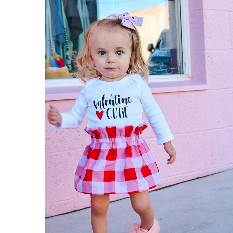 Op de afbeelding: Een jong kind draagt een witte bodysuit met lange mouwen met de tekst "valentine CUTE" in zwarte letters en een rood hart. Het kind draagt ook een rood-witte geruite rok en een roze strik in het haar.