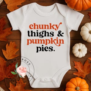 May include: A white short-sleeved baby bodysuit featuring the text "chunky thighs & pumpkin pies." in a mix of orange and black lettering. The design is centered on the chest, and the garment includes three snaps at the bottom for closure.