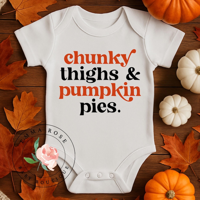 May include: A white short-sleeved baby bodysuit featuring the text "chunky thighs & pumpkin pies." in a mix of orange and black lettering. The design is centered on the chest, and the garment includes three snaps at the bottom for closure.