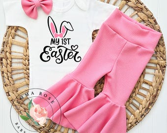 May include: A baby's Easter outfit set. It includes a white onesie with a bunny ear design and the text "My 1st Easter." The set also features pink bell-bottom trousers and a matching pink bow headband. The outfit is displayed on a woven basket.