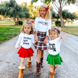 May include: Three children wearing long-sleeved white shirts with black text. One shirt says Christmas cutie, one says better not pout, and one says Santa Baby. The children wear a red skirt, a green skirt, and a plaid skirt, paired with brown boots.