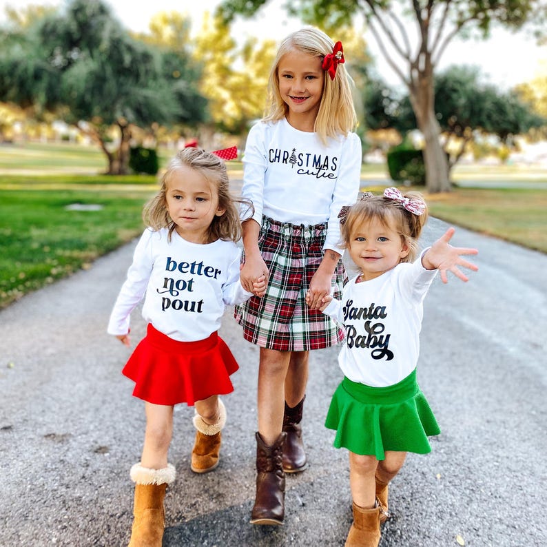 May include: Three children wearing long-sleeved white shirts with black text. One shirt says Christmas cutie, one says better not pout, and one says Santa Baby. The children wear a red skirt, a green skirt, and a plaid skirt, paired with brown boots.