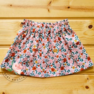 Op de afbeelding: Een rok met bloemenprint en een gerimpelde tailleband. De rok heeft een witte achtergrond met een patroon van roze, oranje en gele bloemen met groene bladeren. De tekst "ROSE'S BABY BOUTIQ" is zichtbaar.