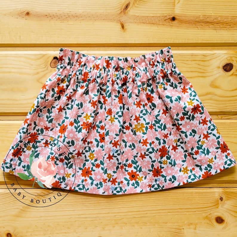 Op de afbeelding: Een rok met bloemenprint en een gerimpelde tailleband. De rok heeft een witte achtergrond met een patroon van roze, oranje en gele bloemen met groene bladeren. De tekst "ROSE'S BABY BOUTIQ" is zichtbaar.