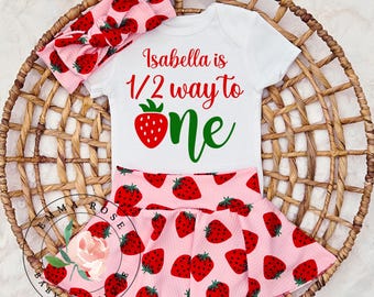 May include: A white short-sleeved bodysuit with the red and green text Isabella is 1/2 way to one and a strawberry graphic, paired with a pink ribbed skirt and a matching headband, both featuring a repeating red strawberry pattern.