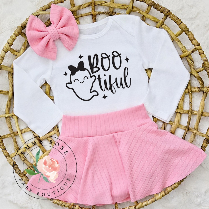 May include: A white long-sleeve bodysuit featuring a black graphic of a ghost with a bow and the text BOO-tiful, paired with a pink ribbed skirt and a matching pink ribbed hair bow. The set includes a circular logo with a floral illustration.