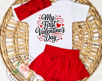 May include: A white baby bodysuit with the text "My First Valentine's Day" in black and red, paired with a red headband and a red skirt. The outfit is displayed on a woven basket.