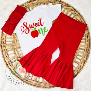 May include: A white short-sleeved bodysuit with the red and green text Sweet One and a red apple graphic, paired with solid red ribbed bell-bottom pants and a matching red ribbed headband with a large bow detail on the front.