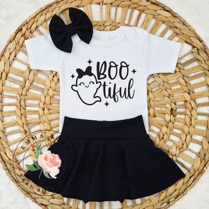 May include: A white short-sleeved bodysuit featuring a black graphic of a ghost with a bow and the text BOO tiful, paired with a solid black skirt and a matching black bow headband. The design includes small black star accents.