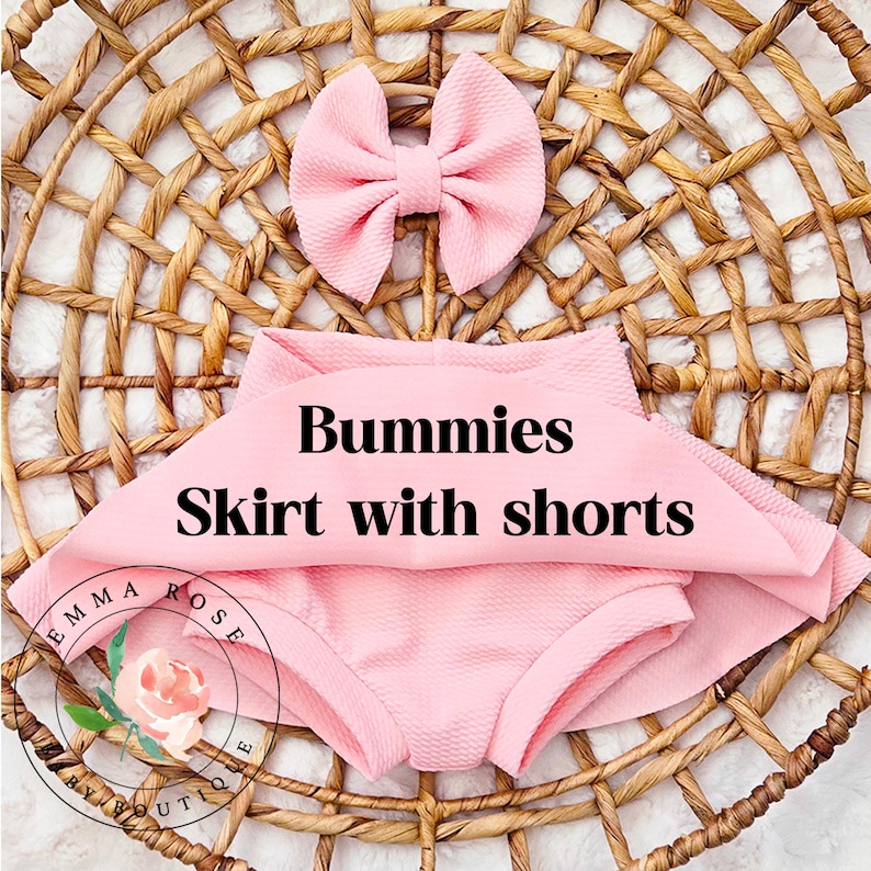 May include: A light pink textured fabric set consisting of a skirt with integrated shorts and a matching hair bow. The text Bummies Skirt with shorts is printed in black across the center. A circular logo with a floral graphic and the text Emma Rose Boutique is visible in the lower left corner.