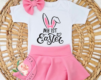 May include: A white baby bodysuit with a black bunny ear graphic and the text "MY 1ST Easter". The outfit includes a pink skirt and a matching pink bow. The outfit is designed for Easter celebrations.