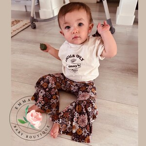 Pu&ograve; includere: Una t-shirt a maniche corte color crema con la scritta LOCALS ONLY e enny's stampata in marrone, abbinata a pantaloni a zampa marroni con un motivo floreale multicolore. La maglietta include una piccola grafica di un cartello stradale.