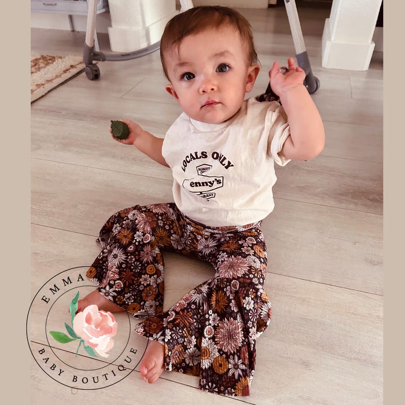 Pu&ograve; includere: Una t-shirt a maniche corte color crema con la scritta LOCALS ONLY e enny's stampata in marrone, abbinata a pantaloni a zampa marroni con un motivo floreale multicolore. La maglietta include una piccola grafica di un cartello stradale.