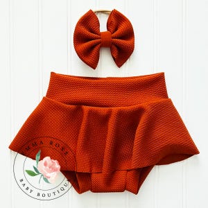 May include: A rust-colored textured fabric bloomer with a flared skirt overlay and a matching bow headband. The set features a consistent deep orange hue and a pebbled surface texture throughout both pieces, presented as a coordinated outfit.