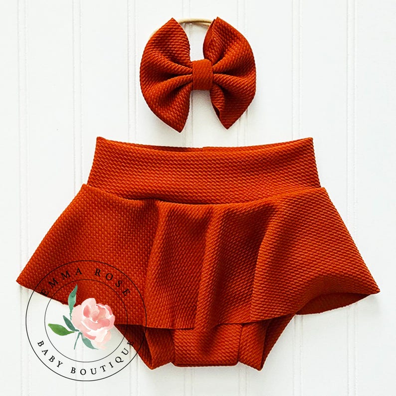 May include: A rust-colored textured fabric bloomer with a flared skirt overlay and a matching bow headband. The set features a consistent deep orange hue and a pebbled surface texture throughout both pieces, presented as a coordinated outfit.
