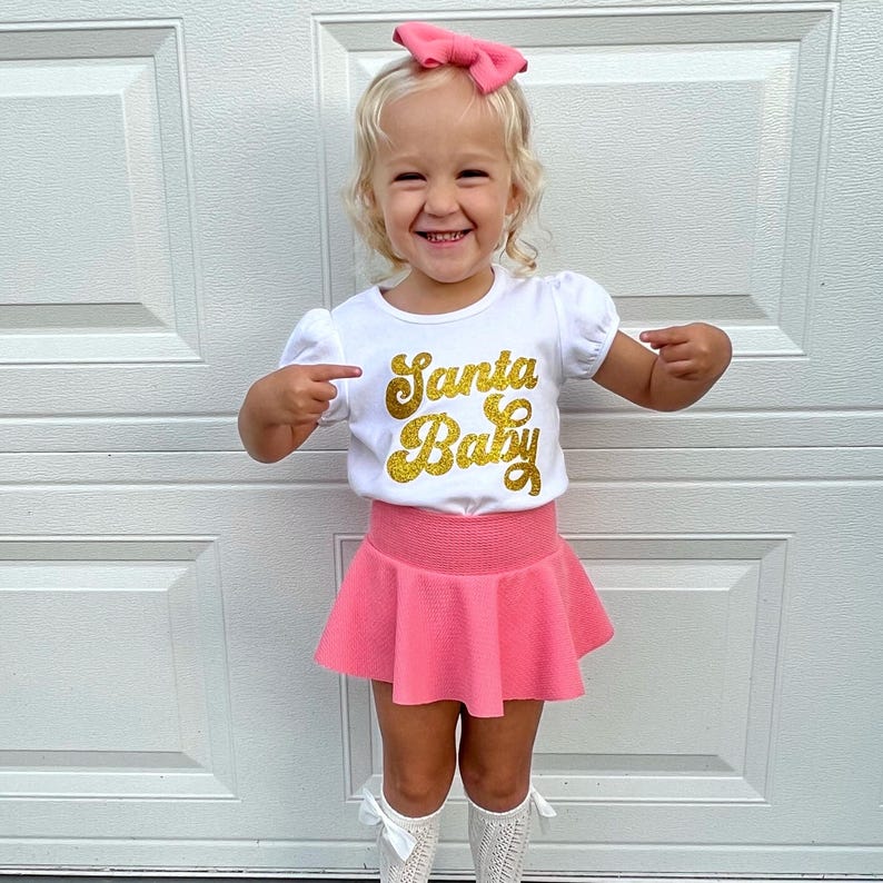 May include: A white short-sleeved t-shirt featuring the text Santa Baby in gold glitter, paired with a pink skirt, a matching pink hair bow, and white textured socks. The shirt has puff sleeves and a round neckline, while the skirt has a flared design.