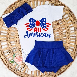 May include: A patriotic baby outfit set featuring a white onesie with a red, white, and blue bow design and the words "All American." The set includes a matching blue headband and a blue skirt. The outfit is displayed on a woven basket.