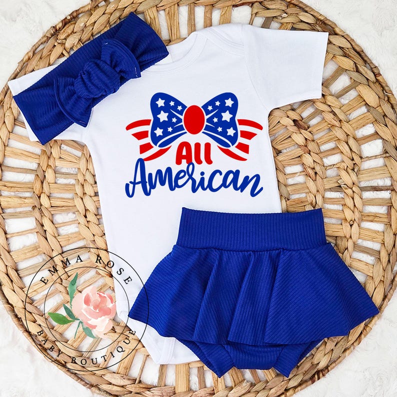 May include: A patriotic baby outfit set featuring a white onesie with a red, white, and blue bow design and the words "All American." The set includes a matching blue headband and a blue skirt. The outfit is displayed on a woven basket.