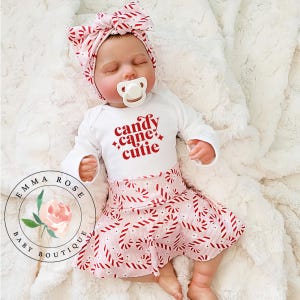 May include: A baby wearing a white long-sleeved onesie with the text "candy cane cutie" in red. The baby is also wearing a pink skirt and matching headband, both with a candy cane pattern. A pacifier is in the baby's mouth.