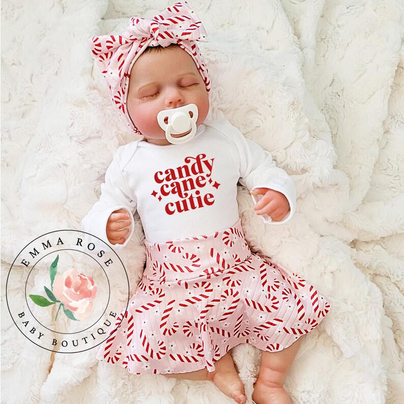 May include: A baby wearing a white long-sleeved onesie with the text "candy cane cutie" in red. The baby is also wearing a pink skirt and matching headband, both with a candy cane pattern. A pacifier is in the baby's mouth.