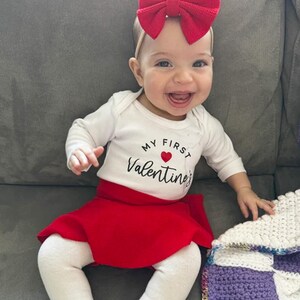 May include: A long-sleeved white bodysuit with the text My First Valentine's and a small red heart printed on the front. The outfit includes a bright red skirt, white leggings, and a large red bow headband worn on the head.