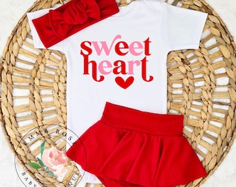 May include: A white short-sleeved bodysuit with the text "sweet heart" in red and pink lettering above a red heart graphic, paired with a red ribbed headband featuring a bow and a matching red ribbed skirted bottom with an elastic waistband.