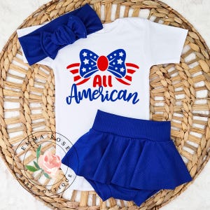 May include: A white baby bodysuit, blue skirt, and matching headband set. The bodysuit features a patriotic design with the words "All American" and a bow with stars and stripes. The skirt and headband are a solid blue color.