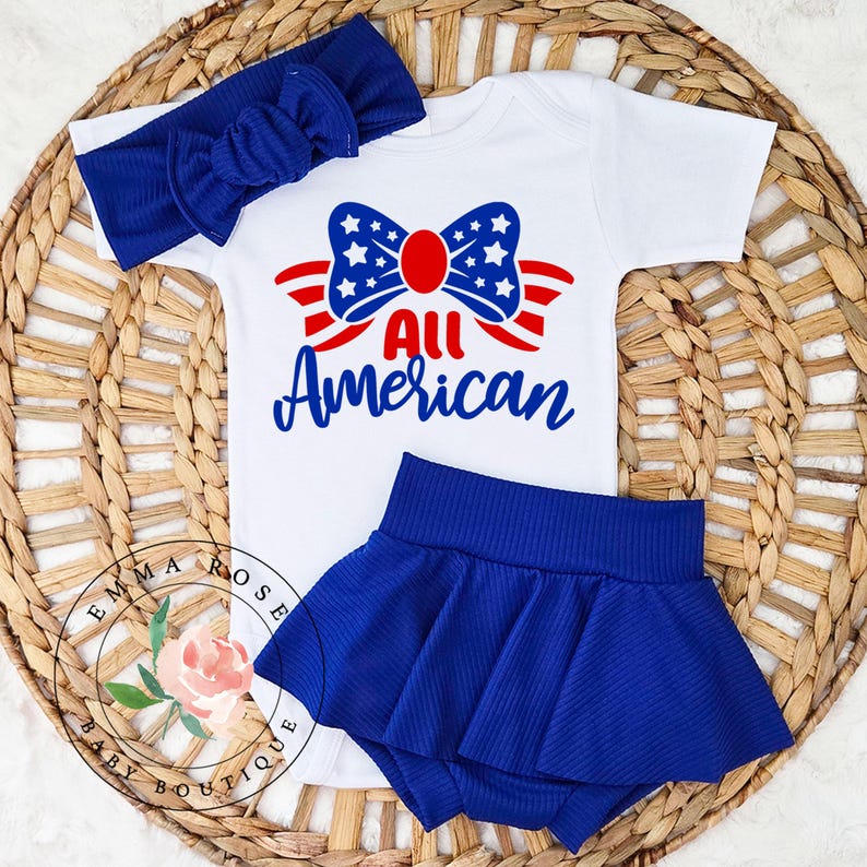 May include: A white baby bodysuit, blue skirt, and matching headband set. The bodysuit features a patriotic design with the words "All American" and a bow with stars and stripes. The skirt and headband are a solid blue color.