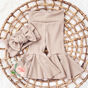 May include: A pair of beige ribbed bell bottom pants with a matching beige ribbed headband. The headband has a large bow. The pants and headband are laid out on a woven basket.