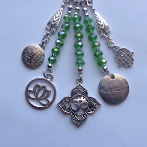 May include: A silver-toned charm cluster featuring a lotus flower, Om symbol, tree of life, Hamsa hand, and a disc with the word "karma." Green faceted beads are strung between the charms. The charms are suspended from a chain.