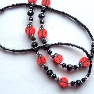 May include: A beaded necklace featuring red ladybug-shaped beads with black spots, interspersed with black cylindrical beads, faceted black round beads, and small silver-toned metallic spacer beads arranged in a repeating pattern.