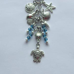 May include: A silver-toned charm bracelet with a beach theme. It features charms of a starfish, seashells, a sand dollar, and a turtle. Blue beads are interspersed throughout the design, adding a pop of color. The charms are connected by small silver rings.