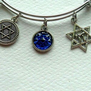 May include: A silver-toned expandable wire bangle bracelet featuring three dangling charms: a circular medallion with a Star of David, a round charm with a faceted deep blue gemstone, and an open-work Star of David charm with a central Hebrew letter.