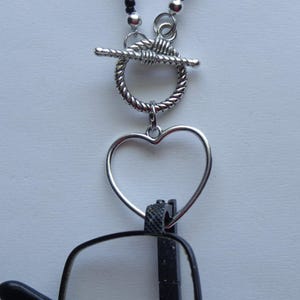 May include: Silver heart-shaped glasses chain with a toggle clasp. The chain is attached to a pair of black eyeglasses.