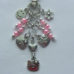 May include: Silver-toned keychain with multiple charms. Charms include a smiling face, flower, pink beaded strands, and several Hello Kitty head pendants. The keychain is attached to a silver clasp.