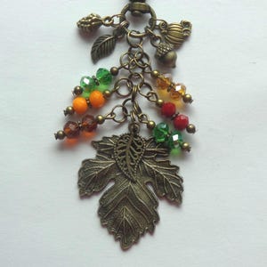 May include: A bronze-toned metal keychain featuring a large maple leaf charm, smaller leaf and acorn charms, and dangling faceted crystal beads in shades of green, orange, red, and amber, all attached to a circular metal ring connector.