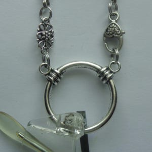 May include: Silver-toned eyeglass holder necklace. The necklace features a large circular pendant with a smaller floral design and a heart-shaped clasp. The glasses are held in place by the circular pendant.